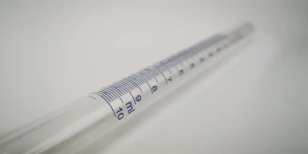 Une image en gros plan montre une pipette transparente avec des graduations en millilitres bleues de 0 à 10, rappelant la précision observée dans les techniques de gravure. La pipette repose sur une surface blanche, sa clarté étant renforcée par un éclairage UV qui met en valeur les mesures détaillées.