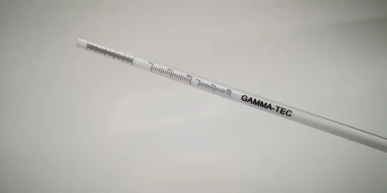 Une sonde blanche avec des repères de mesure et le texte « GAMMA-TEC » imprimé dessus, inclinée en diagonale sur un fond uni, suggérant une précision proche des techniques UV de gravure sur verre.