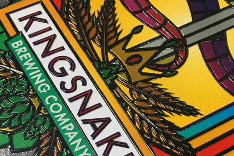 Gros plan dune étiquette de bière colorée et en relief, réalisée selon la technique de limprimante sur objet, comportant linscription « Kingsnake Brewing Company » en texte gras, ornée de tiges de blé, dune couronne et dun fond décoratif qui met en valeur des détails complexes et des couleurs vives.