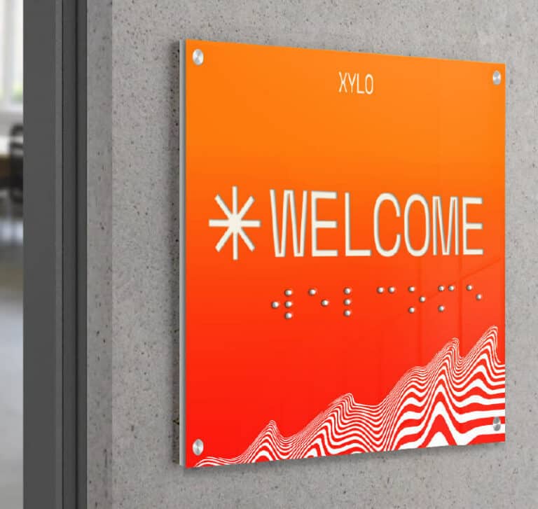 Un panneau orange vif sur lequel est inscrit le mot WELCOME en lettres capitales blanches et points en braille, réalisé à laide de la technologie dimpression sur objet. La partie inférieure présente un motif à rayures rouges et blanches ondulées, avec le mot XYLO élégamment imprimé en haut.