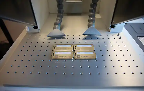 Quatre clés USB dorées étiquetées « Epilog » sont soigneusement placées sur une surface métallique perforée à lintérieur dune machine, probablement en cours de gravure au laser ou dun processus similaire. Deux tuyaux déchappement gris sont stratégiquement positionnés au-dessus delles pour une manipulation optimale.