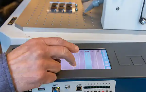 Une personne interagit avec un écran tactile sur un panneau de commande, éventuellement celui d'une machine ou d'un appareil, doté de la technologie à fibre laser. Le panneau comprend des ports USB et réseau, ainsi qu'une série de boutons et d'interrupteurs pour un contrôle galvo précis.