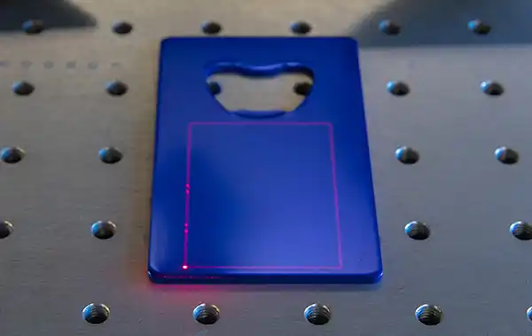 Une feuille de métal bleue au contour rectangulaire repose sur une surface pointillée, avec une découpe laser ressemblant à un décapsuleur sur le dessus. Une grille laser rouge pâle, alimentée par une technologie galvo avancée, est délicatement projetée dessus.