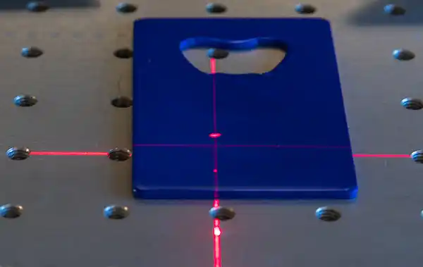 Un objet rectangulaire bleu avec une découpe sur la partie du haut est placé sur une surface métallique avec une grille de trous. Des lignes laser rouges, guidées par des fibres optiques précises, se croisent, formant un signe plus qui s'aligne parfaitement avec le centre de l'objet.