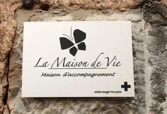 Un panneau sur un mur de pierre indique « La Maison de Vie, Maison daccompagnement » avec un graphique de papillon au-dessus du texte et le logo de la Croix-Rouge française dans le coin.