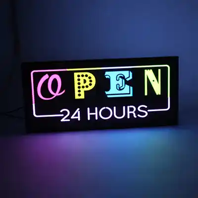 Une enseigne néon rectangulaire avec le mot OPEN en lettres stylisées et 24 HOURS en dessous, s'illuminant en rose, jaune, bleu et blanc sur un fond sombre.