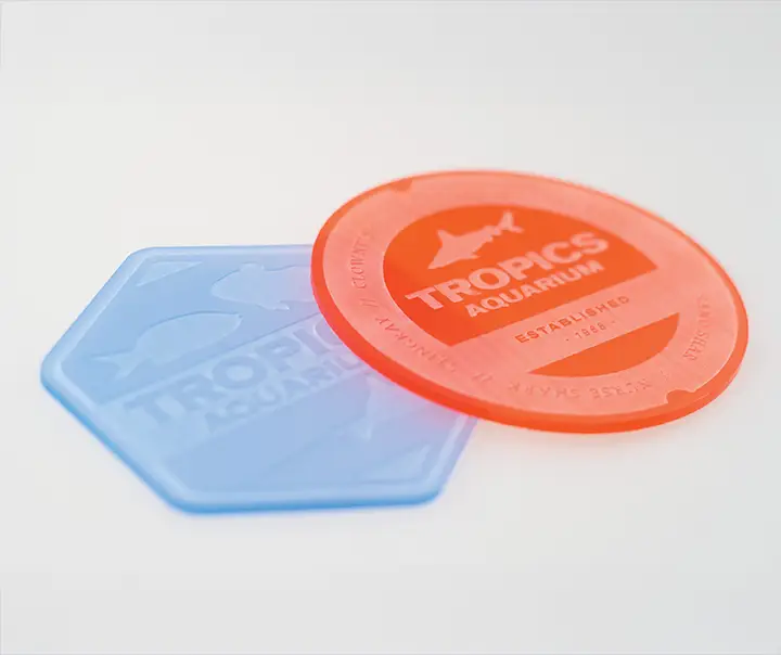Un sous-verre hexagonal bleu translucide et un sous-verre rond orange translucide avec le texte Tropics Aquarium et des graphiques de poissons sont placés sur une surface blanche.