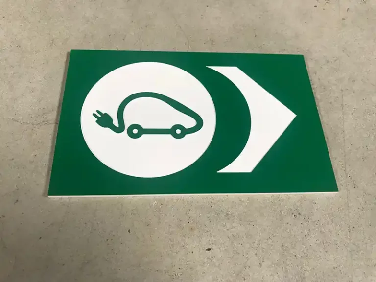 Un panneau vert sur une surface en béton affiche licône dune voiture avec une prise, indiquant la recharge ou le stationnement dun véhicule électrique. Une flèche pointe vers la droite.