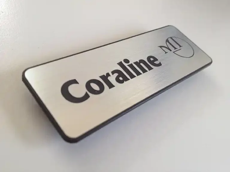 Gros plan dune étiquette nominative argentée avec le mot « Coraline » imprimé en lettres noires en gras. À droite, on peut voir un « M » et un « T » stylisés dans un motif en cercle partiel. Létiquette est placée sur une surface blanche.