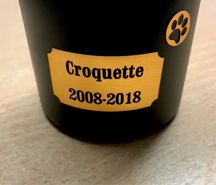 Urne noire avec une étiquette dorée sur laquelle est inscrit Croquette 2008-2018 et un symbole dempreinte de patte au-dessus du texte. Lurne est placée sur une surface en bois.