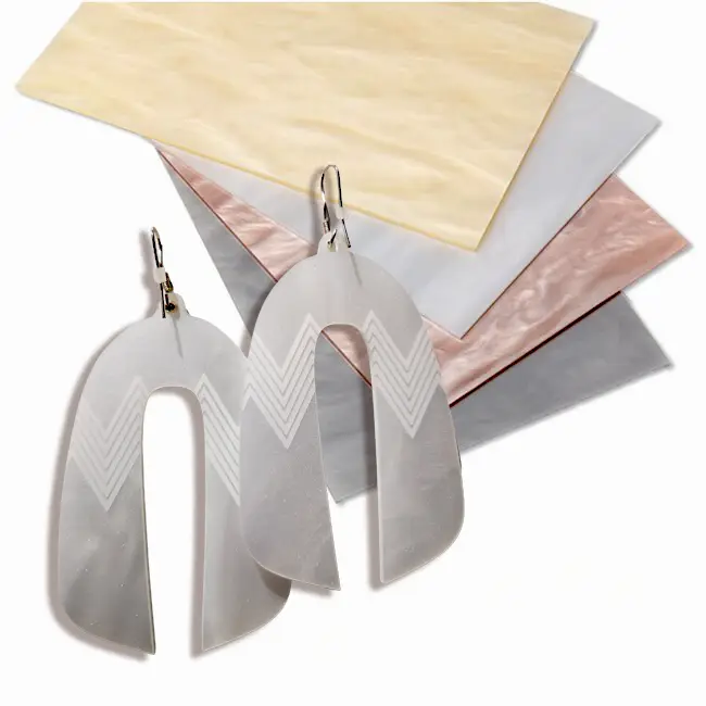 Une paire de boucles d'oreilles translucides en forme d'arche avec des motifs en chevron est présentée devant quatre feuilles rectangulaires d'acrylique marbré de couleur crème, blanche, rose et grise.