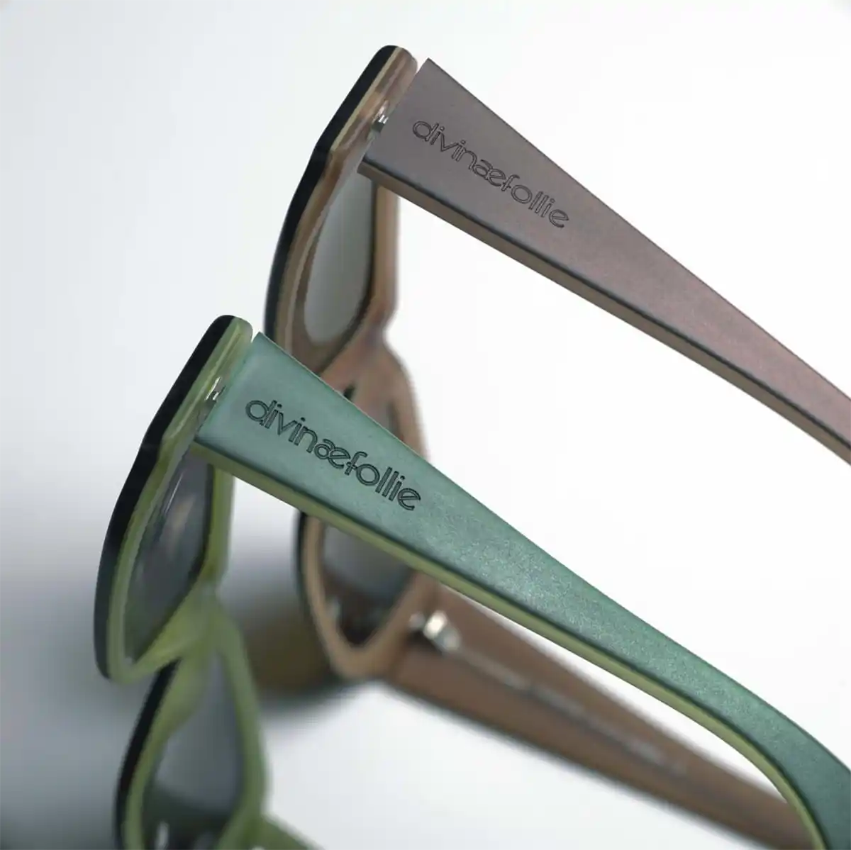 Gros plan de deux paires de lunettes de soleil, lune avec une monture vert olive et lautre avec une monture beige, toutes deux ornées du logo divine&follie. Les experts en marquage et découpe pour lindustrie confectionnent ces pièces exquises, positionnées côte à côte dans un angle élégant.