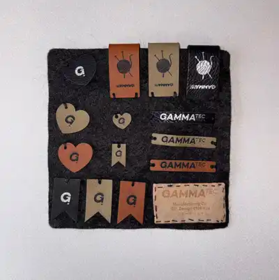 Un tableau carré en feutre noir présentant diverses étiquettes brunes, noires et métalliques de différentes formes, y compris des rectangles, des cercles et des cœurs, comportant pour la plupart la lettre G ou le texte GAMMATEC.