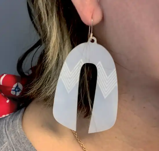 Une personne porte une grande boucle d'oreille en forme de « U » stylisé à l'envers avec des motifs en zigzag. La boucle d'oreille est translucide et est suspendue à un anneau. La personne a les cheveux courts, châtain clair avec des mèches blondes.
