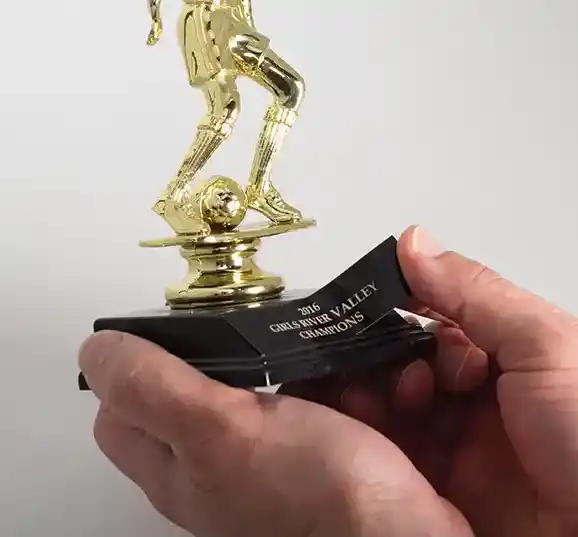 Une personne tient un trophée de football en or avec un joueur en train de dribbler sur une base. La plaque indique « 2014 CHURCHES-IN-VALLEY CHAMPIONS ». Le fond est blanc uni.
