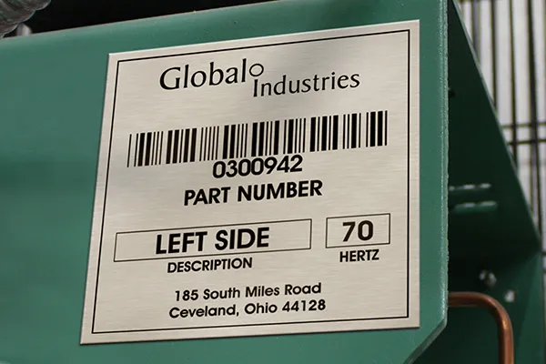 Une étiquette métallique sur l'équipement vert affiche un code-barres, le numéro de pièce 0300942, la description LEFT SIDE, 70 hertz, et l'adresse 185 South Miles Road, Cleveland, Ohio 44128, pour Global Industries.