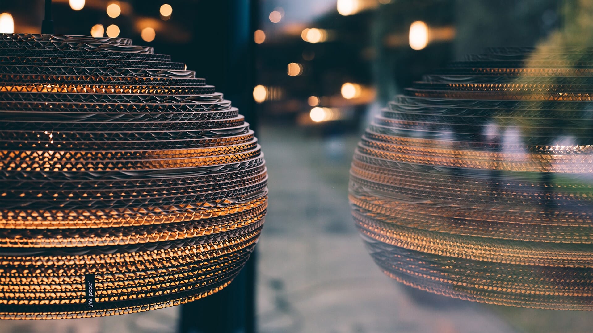 Gros plan dune suspension sphérique texturée de Think Paper avec un éclairage chaleureux, se reflétant sur une fenêtre floue. Larrière-plan présente un doux effet bokeh de lumières supplémentaires, créant une ambiance chaleureuse.
