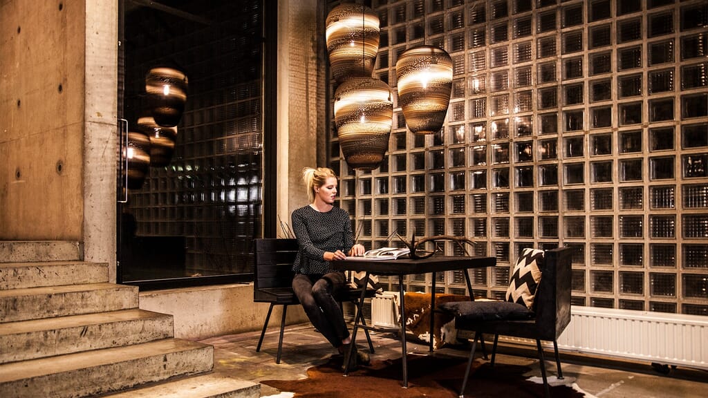 Une personne est assise à une table moderne avec des chaises à motifs, plongée dans un livre, tandis que de grandes lumières artistiques pendent au-dessus de la pièce faiblement éclairée. Le décor respire lambiance Think Paper, avec son mur en blocs de verre texturés et les escaliers et le mur en béton visibles à gauche.