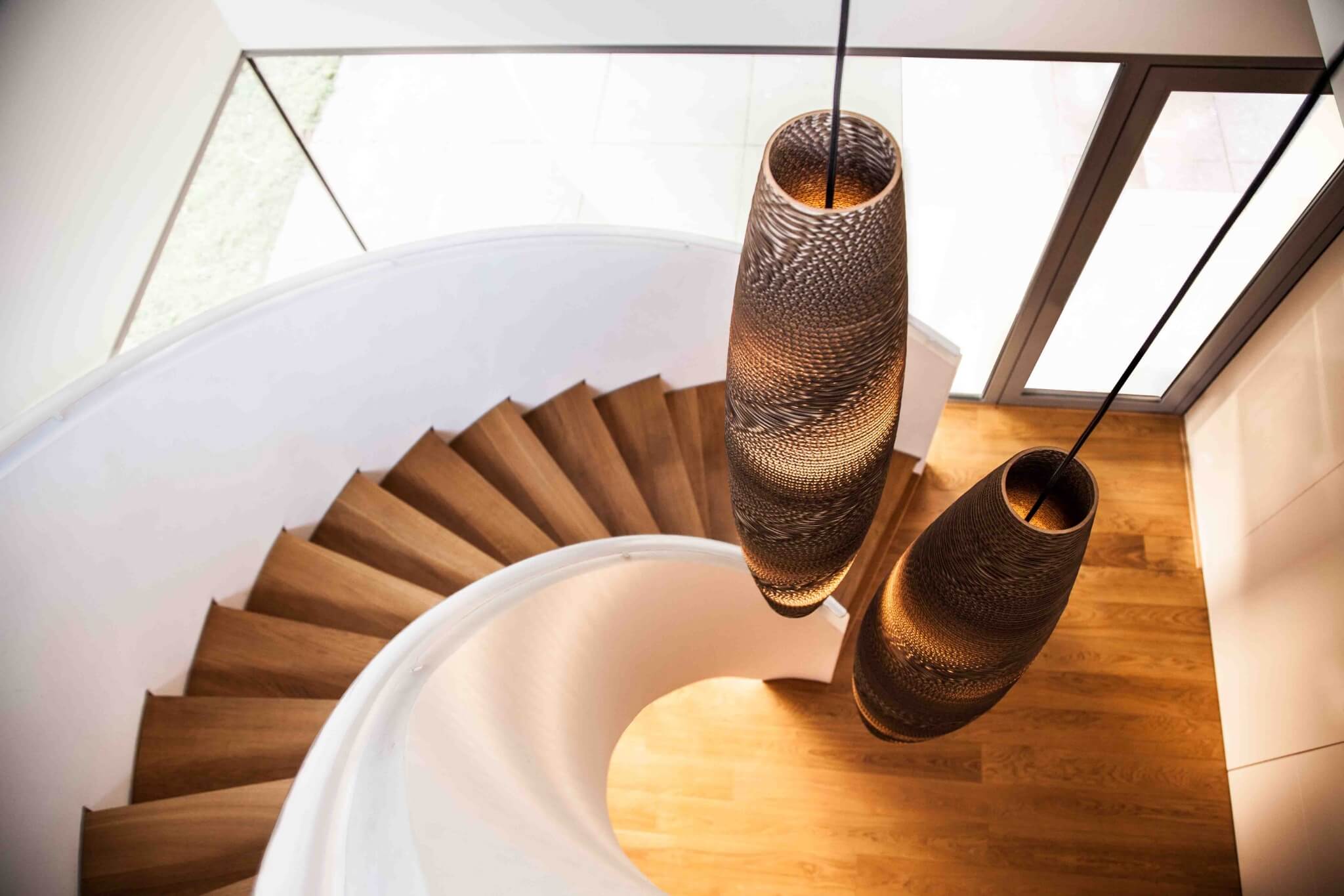 Un escalier en colimaçon avec des marches en bois mène à un espace lumineux. Deux grandes suspensions au design complexe sont suspendues au-dessus, projetant une lumière chaleureuse sur les escaliers. De grandes fenêtres offrent une vue sur lextérieur, ajoutant une lumière naturelle à lespace qui semble aussi inspirante que les créations de Think Paper.