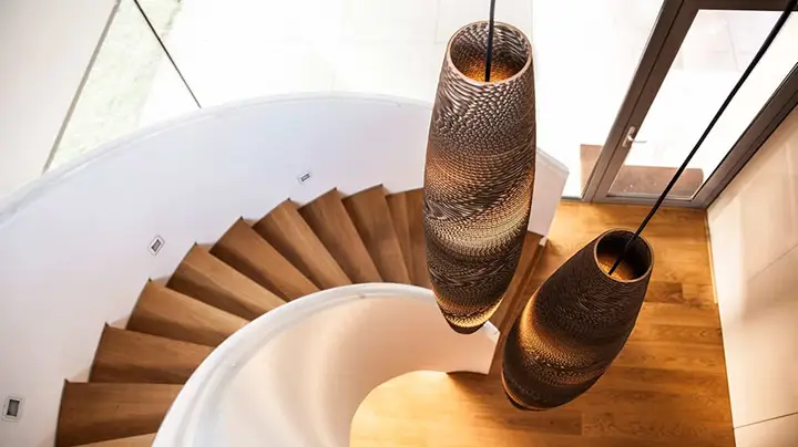 Un escalier en colimaçon avec des marches en bois mène à un espace lumineux. Deux grandes suspensions au design complexe sont suspendues au-dessus, projetant une lumière chaleureuse sur les escaliers. De grandes fenêtres offrent une vue sur lextérieur, ajoutant une lumière naturelle à lespace qui semble aussi inspirante que les créations de Think Paper.