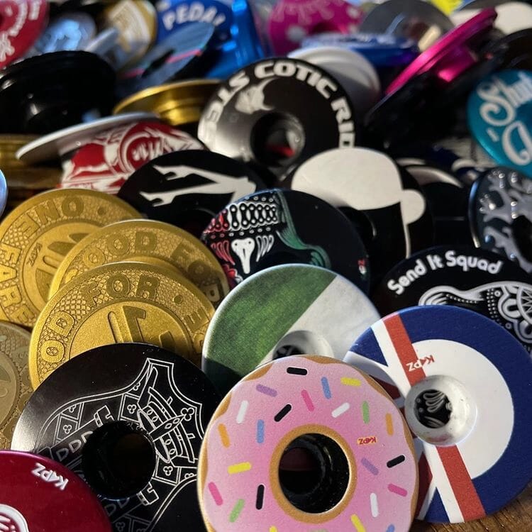 Un assortiment coloré de capsules de bouteilles rondes présente différents motifs, notamment des logos KAPZ, des motifs complexes et des illustrations vibrantes comme un beignet rose et des motifs rayés. Parmi eux se trouve un motif de lotus captivant. Les capsules sont empilées les unes sur les autres, présentant une délicieuse variété de styles.