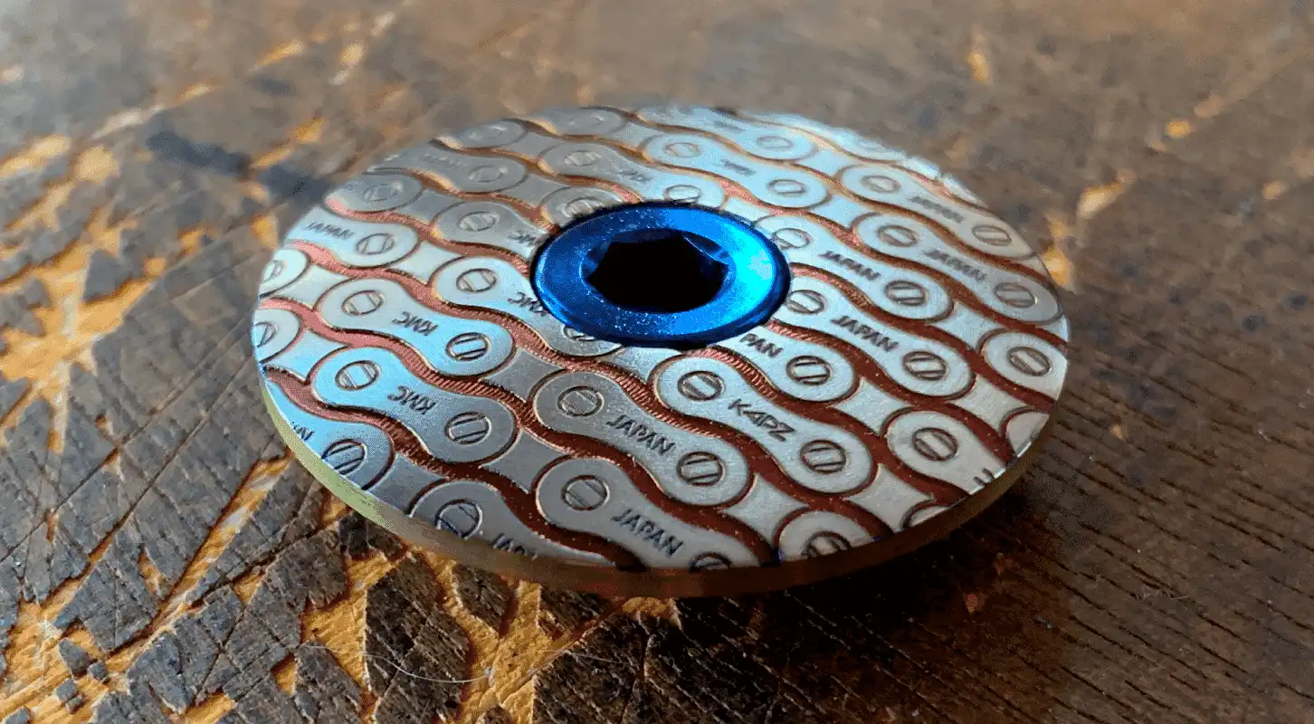 Gros plan dun objet métallique circulaire dont le motif rappelle les maillons dune chaîne de vélo. Le mot « JAPAN » et le logo KAPZ sont représentés en répétition. Louverture centrale hexagonale bleue rappelle une fleur de lotus. Larrière-plan est une surface en bois texturée.