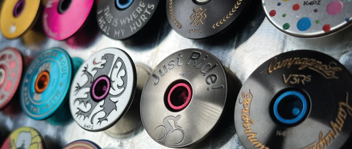 Gros plan de divers capuchons de casque de vélo colorés, dont un modèle KAPZ en forme de lotus, montés sur une surface métallique. Les capuchons présentent différents motifs, symboles et textes tels que « Just Ride ! » ainsi que des motifs décoratifs. Ils ont de petits trous centraux pour le montage.