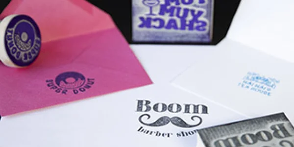 Une collection de timbres en bois, fabriqués avec une précision proche du travail à la machine, et des enveloppes sur une surface sombre. Les timbres présentent des motifs tels que « MUY MUY SHACK » et « Boom barber shop » avec une moustache. Les enveloppes en dessous affichent fièrement des empreintes en violet et bleu.