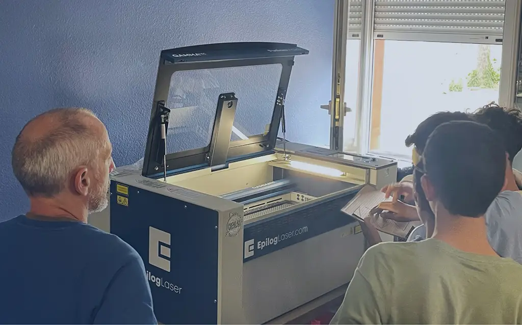 Trois personnes se tiennent devant une machine Epilog Laser ouverte, mettant en évidence l'enseignement pratique du laser. Une personne tient des documents tandis qu'une quatrième fait face au groupe dans une salle aux murs bleus où la lumière du jour passe par une fenêtre ouverte.