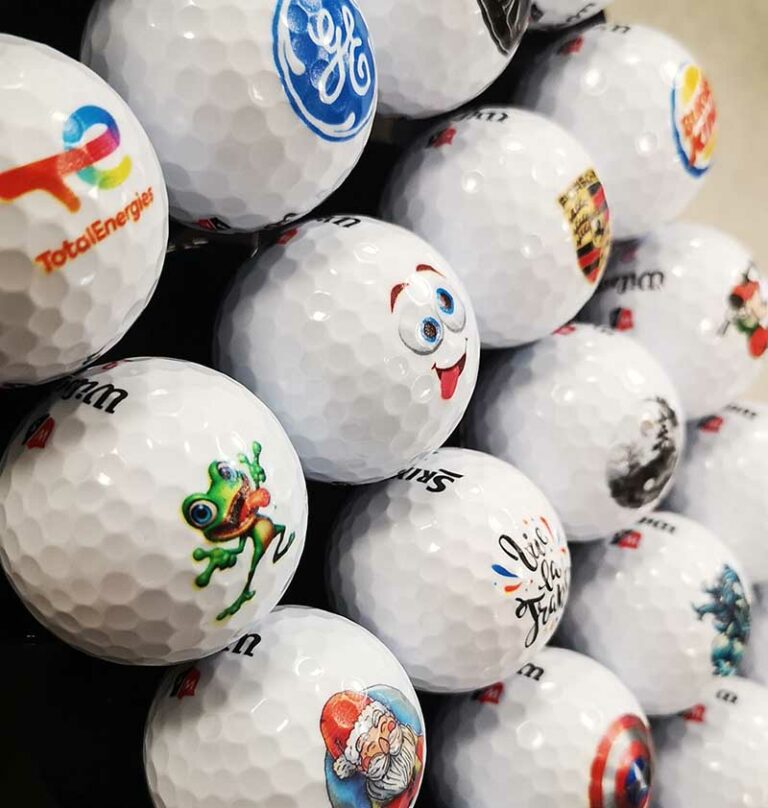 Gros plan sur des balles de golf blanches, chacune ornée d'un logo coloré différent ou d'un dessin humoristique créé à l'aide d'une Imprimante Roland, disposées dans un présentoir bien ordonné.