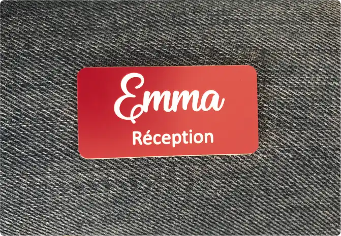 Une étiquette nominative rouge avec le texte « Emma Réception » écrit en blanc, posée sur un fond texturé foncé.