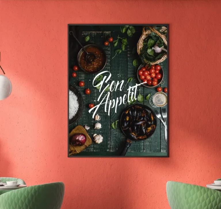 Une image encadrée sur un mur rouge - créée avec l'imprimante sur objet - montre des ingrédients et des plats sur une surface en bois vert foncé avec le texte Bon Appétit en écriture blanche. Deux chaises vertes et des lampes suspendues blanches sont partiellement visibles.