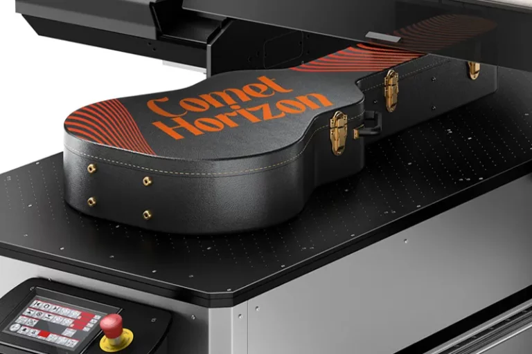 Un étui de guitare portant le texte Comet Horizon en orange est placé à l'intérieur d'une grande imprimante sur objet, dont la surface semble imprimée. L'étui noir est doté d'une quincaillerie dorée et d'accents rayés rouges.