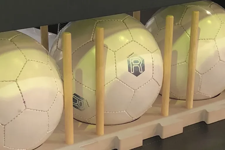 Plusieurs ballons de football blancs avec des panneaux cousus et un logo R imprimé, créé à l'aide d'une imprimante sur objet, sont rangés derrière des chevilles verticales en bois sur une étagère de couleur claire.