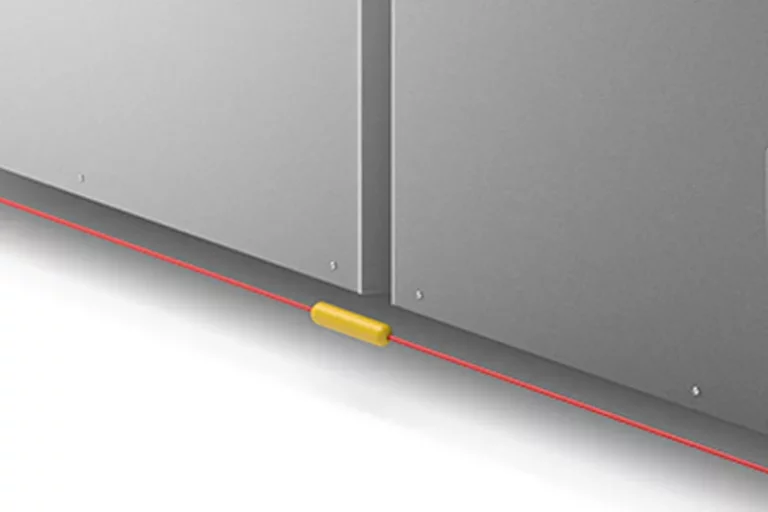 Un capteur jaune est monté près de la base d'un panneau métallique gris. Une ligne laser rouge, souvent utilisée avec une imprimante sur objet pour un alignement précis, court horizontalement le long du sol, correspondant au capteur et à la base du panneau.