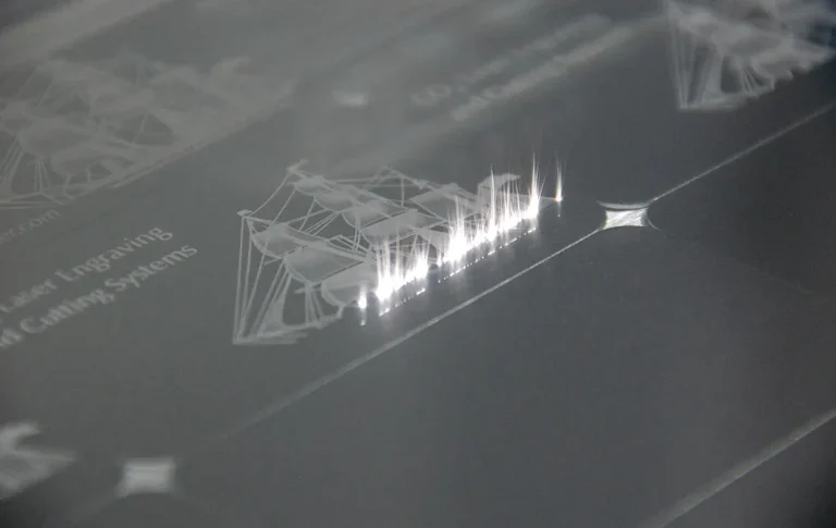 Gros plan d'une machine de gravure laser gamma tec gravant une illustration de bateau et du texte sur un matériau plat et transparent, avec des étincelles lumineuses visibles à l'endroit où le laser coupe.