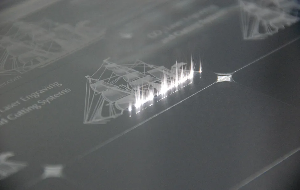 Gros plan d'une machine de gravure laser gamma tec gravant une illustration de bateau et du texte sur un matériau plat et transparent, avec des étincelles lumineuses visibles à l'endroit où le laser coupe.