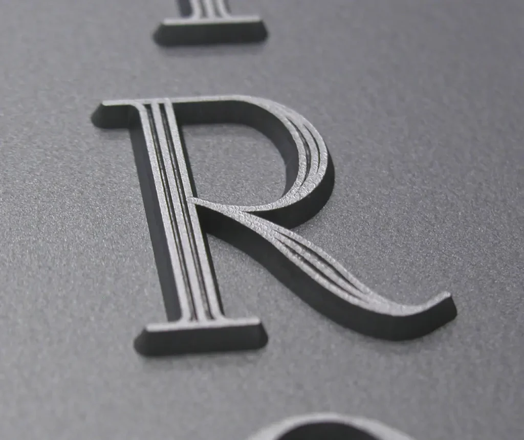 Gros plan d'une lettre R en relief et en relief avec des lignes parallèles gravées sur une surface lisse, gris foncé. L'image met l'accent sur la texture et les détails de la lettre.