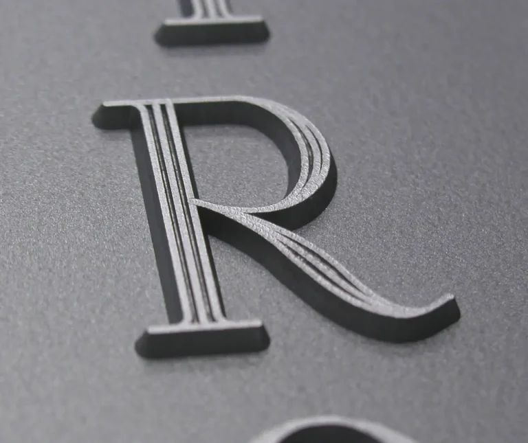 Gros plan d'une lettre R en relief et en relief avec des lignes parallèles gravées sur une surface lisse, gris foncé. L'image met l'accent sur la texture et les détails de la lettre.