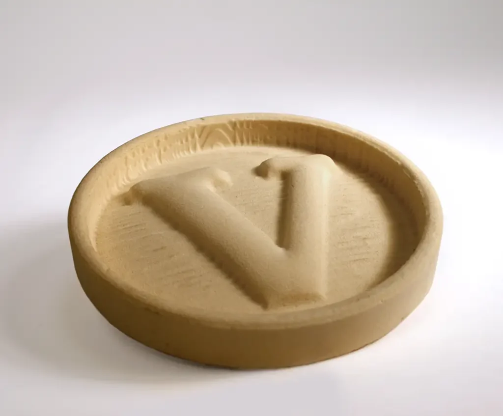 Un plateau rond de couleur beige avec la lettre V en relief au centre, fabriqué dans un matériau qui ressemble à du sable ou à de la poudre comprimée, placé sur une surface blanche unie.