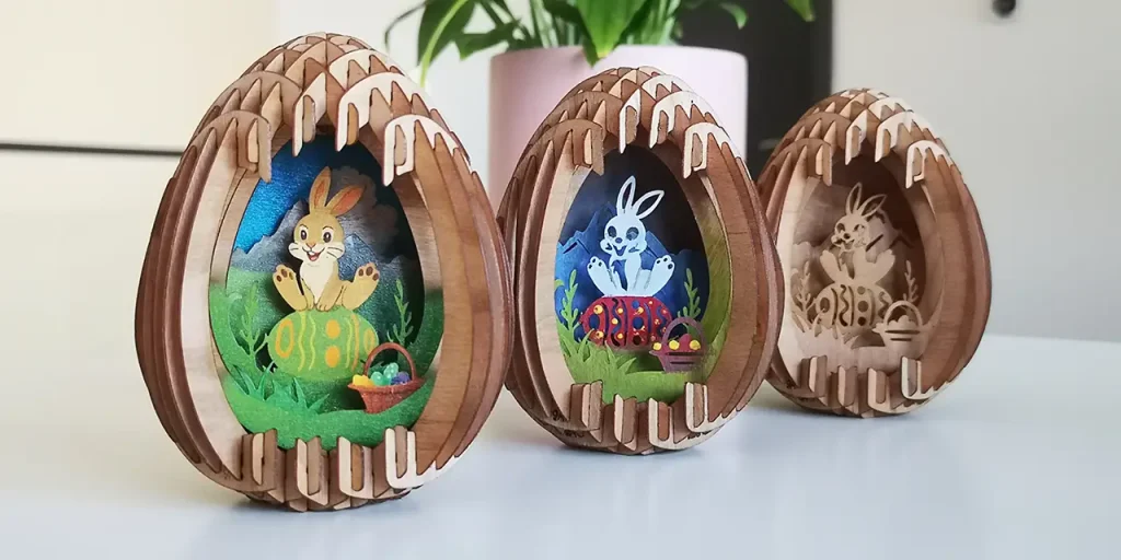 Trois ornements en bois en forme d'œuf avec des motifs complexes de découpe de pâques au laser, représentant des lapins de Pâques et des œufs décorés, placés sur une surface blanche avec une plante en pot à l'arrière-plan.