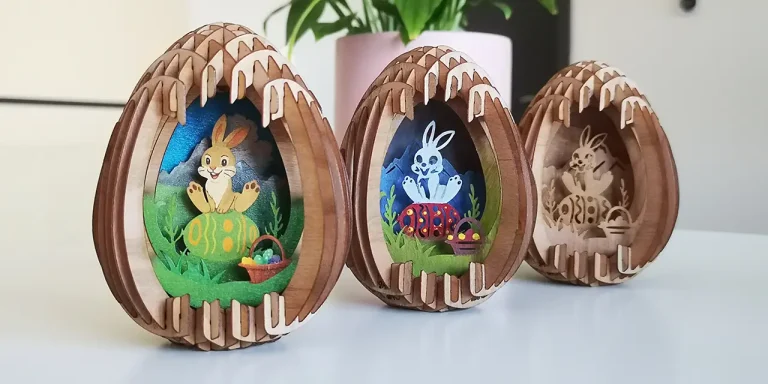 Trois ornements en bois en forme d'œuf avec des motifs complexes de découpe de pâques au laser, représentant des lapins de Pâques et des œufs décorés, placés sur une surface blanche avec une plante en pot à l'arrière-plan.