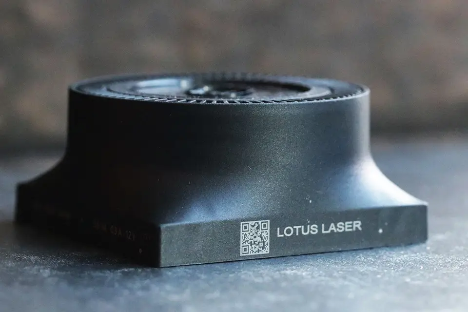 Un objet métallique cylindrique noir avec une large base, marqué "LOTUS LASER" à côté d'un code QR, démontre le marquage industriel sur une surface sombre.