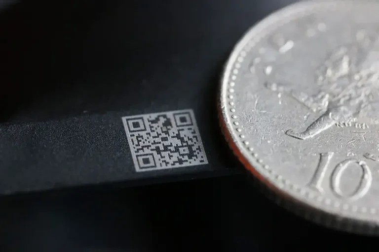 Une image en gros plan montrant un petit code QR imprimé à côté de la tranche d'une pièce de monnaie, mettant en évidence la précision du marquage industriel dans la production de codes minuscules pour l'identification des produits.