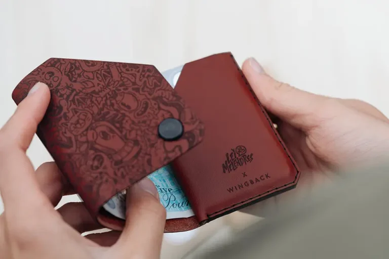Une personne tient un portefeuille ouvert en cuir marron avec un motif en relief, présentant le marquage industriel et les mots Wingback et Memento Mori à l'intérieur. Des billets de banque et des cartes sont partiellement visibles dans le portefeuille.