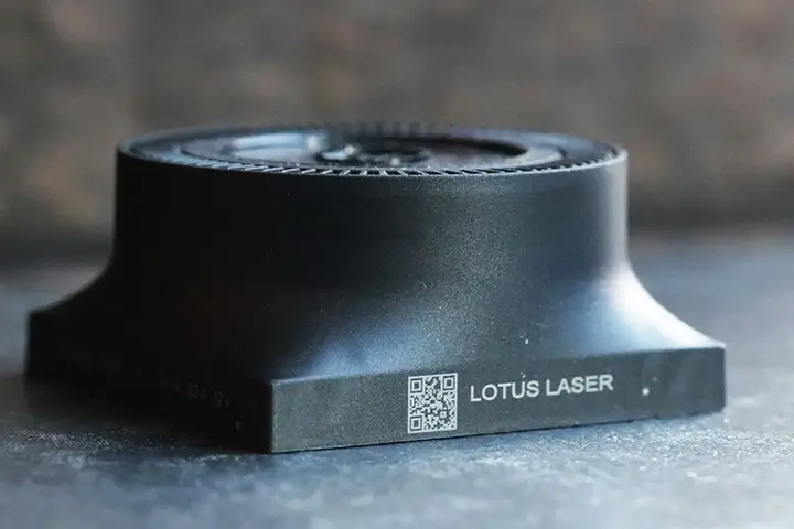 Un objet cylindrique noir avec une large base est représenté sur une surface. L'objet présente un marquage industriel, dont LOTUS LASER et un code QR gravé sur le côté. L'arrière-plan est flou.