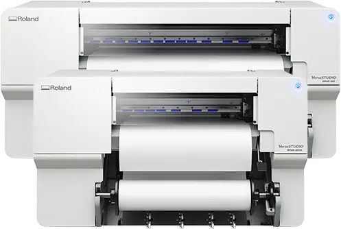 Deux imprimantes blanches Roland VersaSTUDIO imprimante/découpeuse jet d'encre sont placées l'une derrière l'autre, toutes deux chargées de rouleaux de papier vierge. Ces machines modernes affichent des voyants bleus et sont prêtes à effectuer des tâches d'impression et de découpe précises.