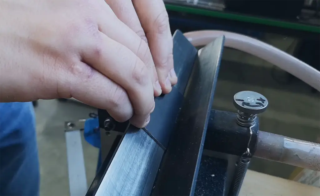 Une personne utilise ses mains pour affûter le bord d'un ski sur un outil en métal dans un atelier de mécanique, avec une vis et un tuyau visibles à l'arrière-plan.