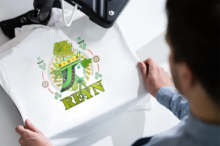 Une personne utilise une presse à chaud pour appliquer un graphique coloré d'une reine avec le mot REYN sur un T-shirt blanc, créé avec une Imprimante Roland. Ses mains maintiennent le tee-shirt en place sous la machine.