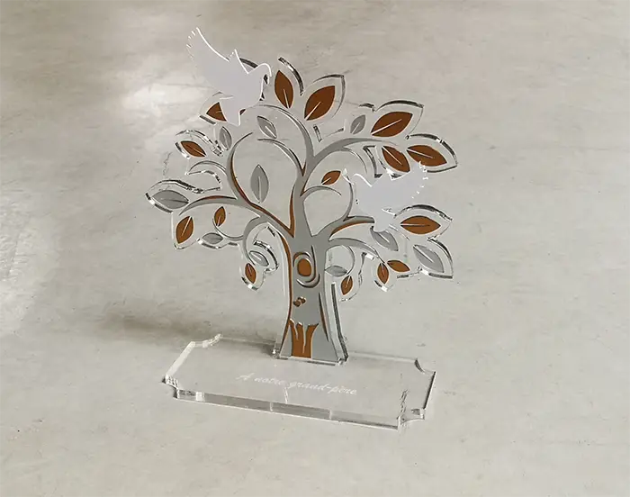 Un trophée en acrylique transparent en forme d'arbre avec des feuilles découpées et deux colombes blanches, réalisé avec un laser découpeur, repose sur une base rectangulaire. Le fond est une surface lisse de couleur claire.
